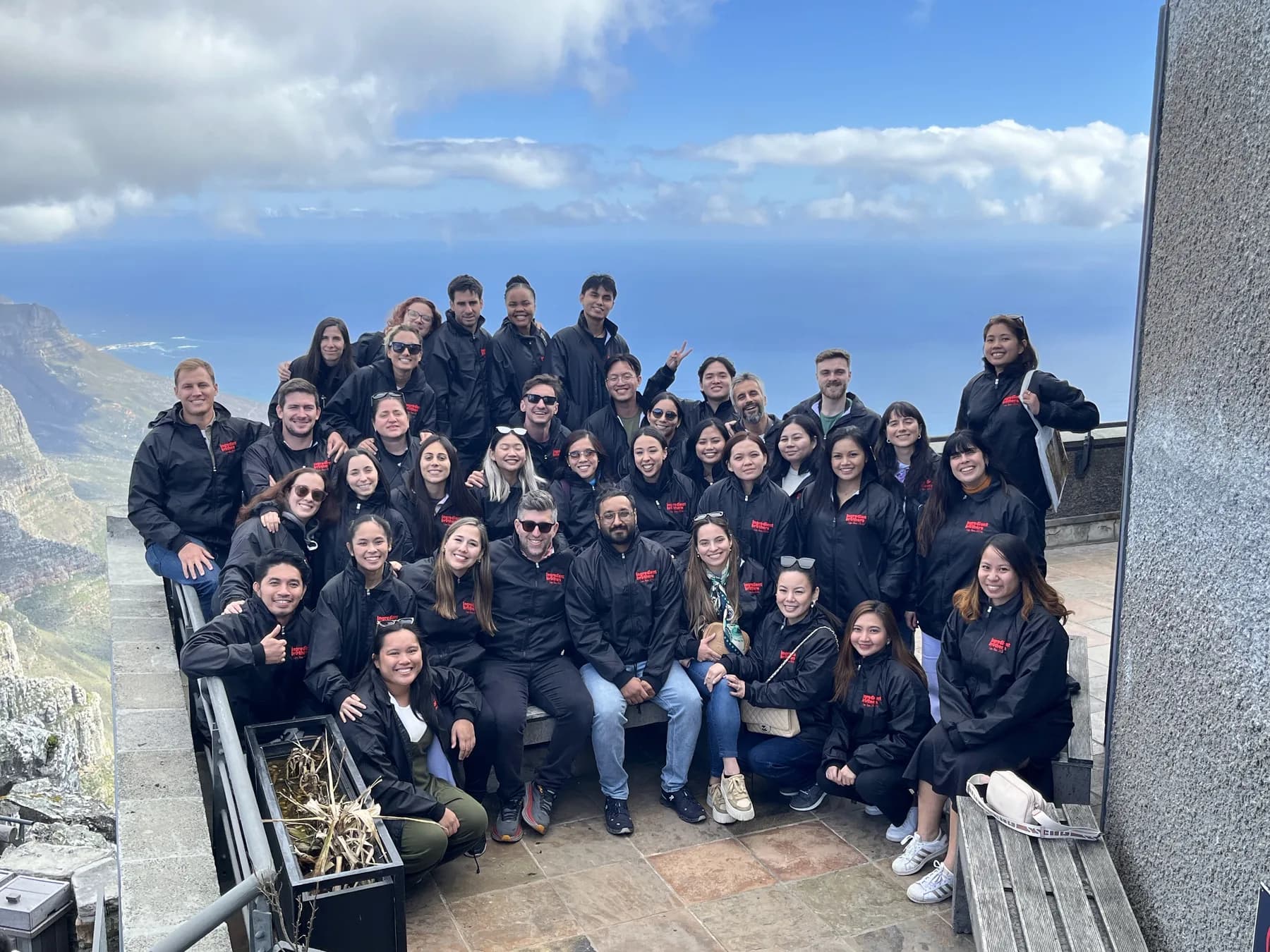 The team in matching black jackets at a mountaintop lookout above the ocean.